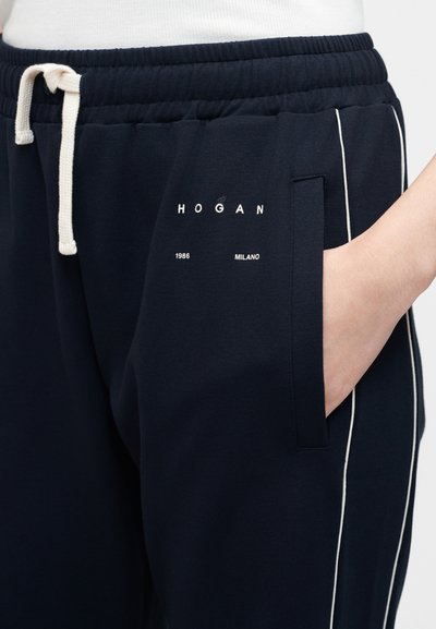 Σκούρες μπλε φόρμες με λευκό κορδόνι, πλαϊνές τσέπες, λευκή ρίγα και το κείμενο "HOGAN 1986 MILANO" στο αριστερό μηρό.