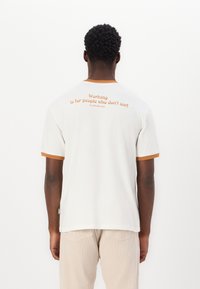 Camiseta blanca de manga corta con cuello y puños acanalados en naranja, que presenta un texto en la parte de atrás: "Trabajar es para personas que no surfean."