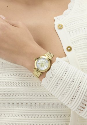 Zlatni ručni sat s okruglim brojčanikom i rimskim brojevima na osobi koja nosi bijelu teksturiranu majicu dugih rukava s zlatnim gumbima.