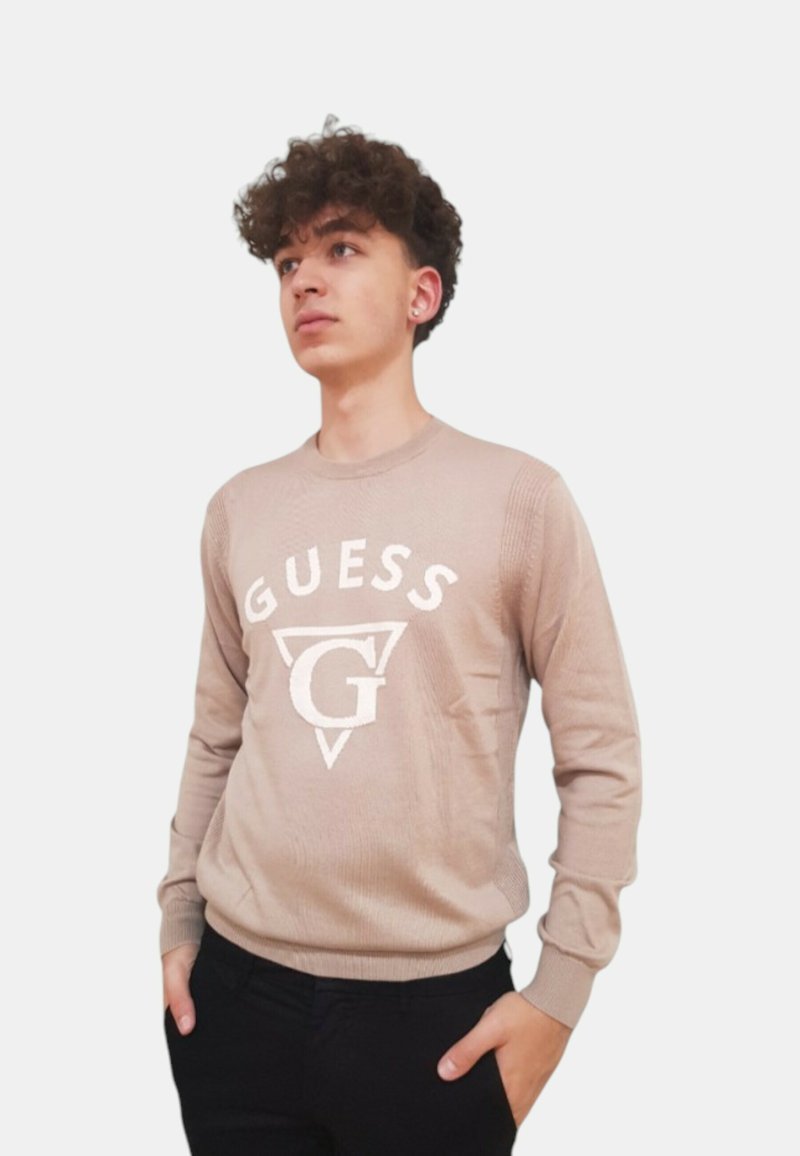 Maglione in maglia beige con logo "GUESS" bianco sul davanti, dotato di scollatura rotonda e polsini a coste, abbinato a pantaloni neri.