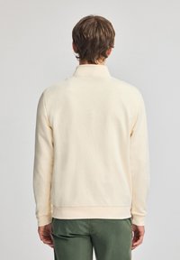 Sudadera color crema con cuello, mangas largas y puños y dobladillo de canalé. Textura suave y diseño minimalista sin logotipos visibles.