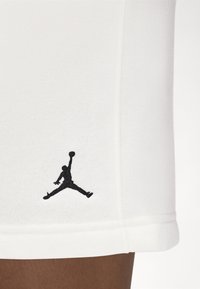Close-up van witte basketbalschoenen met zwart Jumpman-logo vlak boven de zoom, naast een donkergekleurde been.