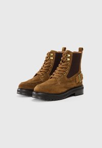 Bocanci din suede maro cu șireturi, panouri elastice negre pe lateral, ochiuri aurii, talpa groasă neagră și catarame decorative pe lateral.