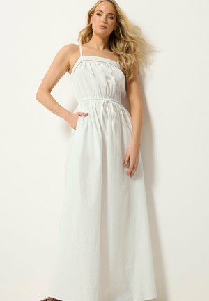 Frau mit langen blonden Haaren, die ein ärmelloses weißes Maxikleid mit Kordelzug an der Taille und Seitentasche trägt, steht vor einem hellen Hintergrund.