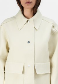 Light beige jacket made of sturdy fabric, featuring a wide collar, two large patch pockets, and metal snap closures. Smooth texture.