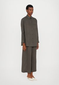 Camicia in lino verde scuro, comoda e con bottoni, dotata di taschino sul petto, abbinata a pantaloni larghi coordinati e scarpe beige a punta.