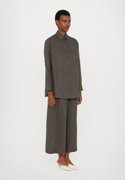 Χαλαρή, σκούρα πράσινη λινή πουκάμισο με κουμπιά και τσέπη στο στήθος, συνδυασμένο με αντίστοιχες φαρδιές παντελόνες και μπεζ μυτερές εσπαντρίγιες.