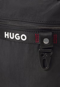 Borsa nera in nylon con zipper visibile, logo "HUGO" bianco, dettagli in cuciture rosse e clip a moschettone nera. Texture liscia e finitura resistente.