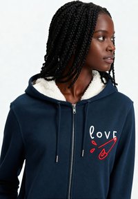 Sweat à capuche zippé navy avec doublure en Sherpa blanche, présentant un design brodé en rouge de "love" et une épingle de sûreté sur la poitrine gauche. Texture lisse.