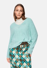 Helltürkiser, flauschiger Pullover mit V-Ausschnitt, über einem weißen Oberteil getragen. Kombiniert mit einem gemusterten Rock in Teal-, Braun- und Cremefarben mit einem Kordelzug.
