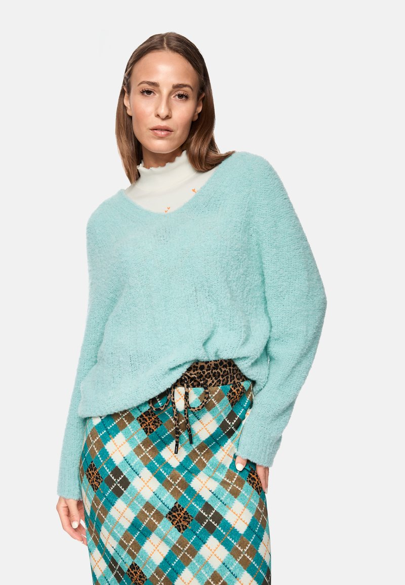 Helltürkiser, flauschiger Pullover mit V-Ausschnitt, über einem weißen Oberteil getragen. Kombiniert mit einem gemusterten Rock in Teal-, Braun- und Cremefarben mit einem Kordelzug.