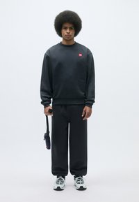 Sudadera negra y pantalones de chándal a juego, hechos de un material suave. La sudadera tiene un pequeño logo rojo; el modelo sostiene un bolso negro de strap.