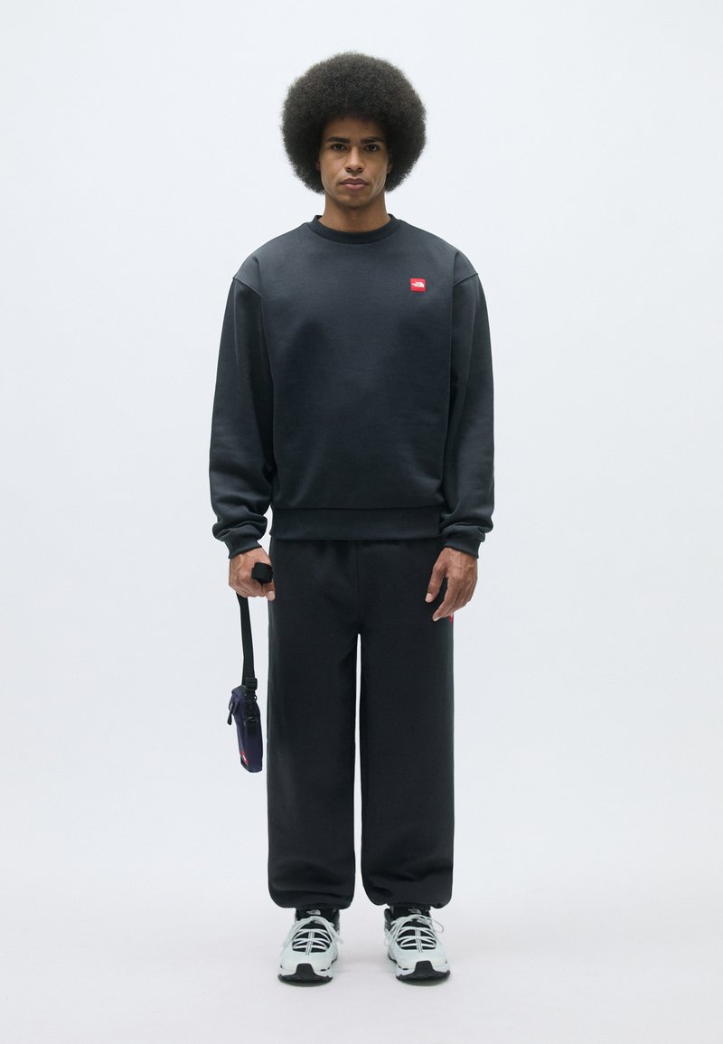Sweatshirt noir et pantalon de jogging assorti, fabriqués en matériau doux. Le sweatshirt présente un petit logo rouge ; le modèle tient un sac bandoulière noir.
