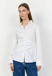 Camicia bianca a maniche lunghe con design avvolgente, chiusura con bottoni davanti e polsini oversize, abbinata a pantaloni in denim blu. Tessuto liscio e fresco.