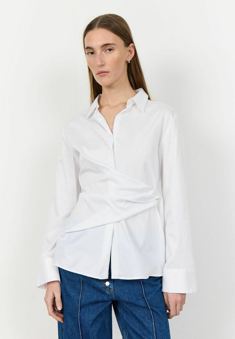 Camicia bianca a maniche lunghe con design avvolgente, chiusura con bottoni davanti e polsini oversize, abbinata a pantaloni in denim blu. Tessuto liscio e fresco.
