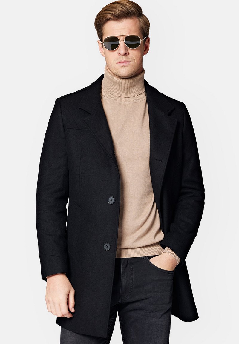 Czarny wełniany płaszcz z klapami i dwoma przednimi guzikami, noszony na beżowym golfie. Posiada elegancki design i nowoczesne okulary przeciwsłoneczne.