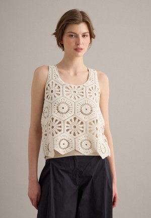 Vrouw draagt een mouwloze, crèmekleurige gehaakte top met zeshoekige bloemmotieven over een beige ondershirt en zwarte broek, staand tegen een effen achtergrond.