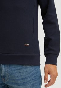 Hanorac bleumarin dintr-un material texturat cu un model în grilă, având manșete și tiv dintr-un material ribbed și o mică etichetă a brandului pe partea de jos.