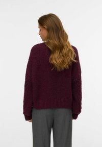 Pull en maille bordeaux avec une texture épaisse et des détails en torsades sur les manches, associé à un pantalon gris. Vu de dos.
