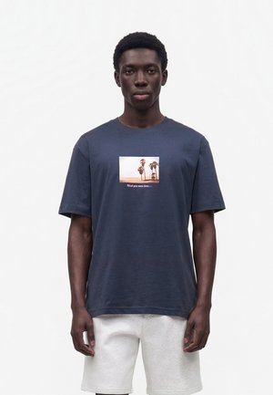 Jeune homme portant un t-shirt bleu marine avec un motif de palmier et du texte, associé à un short gris clair, debout sur un fond blanc uni.