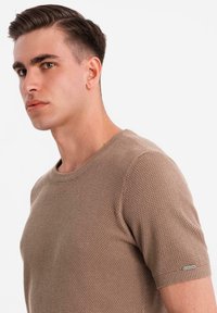 Camisa de punto texturizada beige de manga corta con cuello redondo y un sutil toque metálico en la manga izquierda, llevada por un modelo.