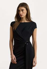 Zwarte jurk met een aansluitende silhouet, kapmouwen en een gedraaide voorzijde. Heeft een metalen ringaccent op de taille. Glad textuur.