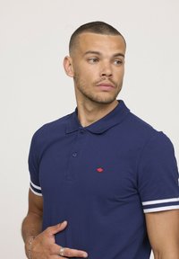 Polo shirt bleu marine avec col, deux boutons et un petit logo rouge. Il présente des rayures blanches sur les manches et est fait d'un tissu texturé.
