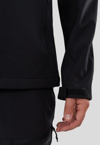Närbild av en svart jackärm med justerbart kardborrefäste på en persons hand mot en grå bakgrund.
