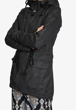 Kobieta w czarnej kurtce z kapturem ze ściągaczami, na suwak z przodu, z dużymi kieszeniami i wzorzystymi spodniami, z rękami skrzyżowanymi na piersiach.
