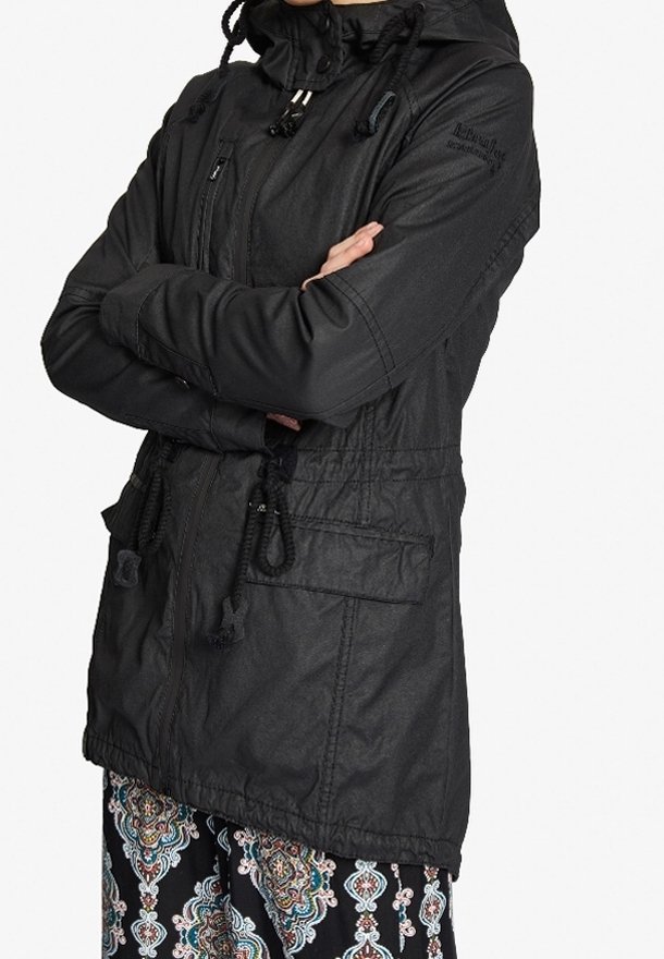 Mujer con chaqueta negra con capucha con cordones, cremallera al frente, bolsillos grandes y pantalones estampados, con los brazos cruzados al frente.