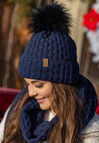 Sötétkék kötött sapka, amelynek tetején egy nagy fekete szőr pom-pom található. A sapka texturált mintával készült, az oldalán pedig egy kis barna bőr címke van.