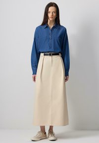 Denim button-up shirt with patch pockets, paired with a cream-colored pleated maxi skirt and beige sneakers. Black belt at the waist.