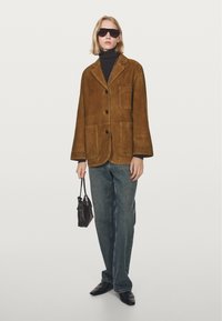 Giacca in suede marrone con tre bottoni, una tasca sul petto e due tasche frontali; indossata sopra un dolcevita nero e abbinata a jeans scuri e scarpe.