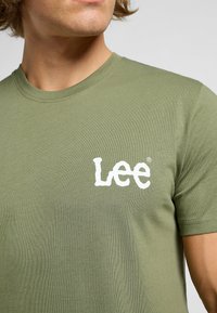 Oliwkowa bawełniana koszulka z okrągłym dekoltem, z białym logo graficznym "Lee" na piersi. Krótkie rękawy i gładka faktura.