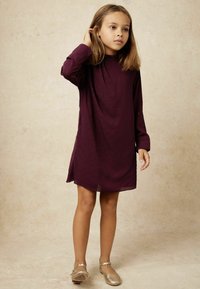 Ein junges Mädchen steht mit einem langärmligen lila Kleid mit kleinen Perlen und goldenen Mary Jane Schuhen vor einem neutralen Hintergrund.