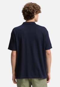 Marineblaues Polohemd mit kurzen Ärmeln, gerippter Textur und klassischem Kragen. Hat einen geraden Schnitt und ein kleines Etikett-Detail am Saum.