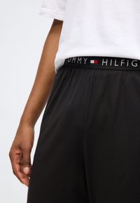 Un pantalon de survêtement noir avec une taille élastique portant le logo "TOMMY HILFIGER" et des accents de couleur contrastés. Le tissu semble doux et lisse.