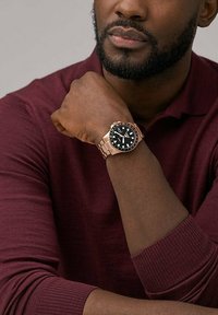 Schwarz Zifferblatt Armbanduhr mit einem Armband aus Roségoldmetall, ausgestattet mit leuchtenden Zeigern, einer geriffelten Lünette und einer strukturierten Oberfläche. Am Handgelenk getragen.