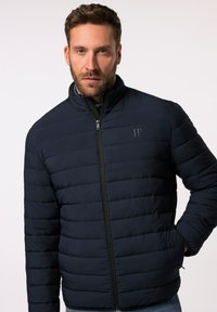 Giacca imbottita blu navy con colletto alto, chiusura con zip e cuciture verticali. Logo sul lato sinistro. Tessuto liscio con una vestibilità moderna.