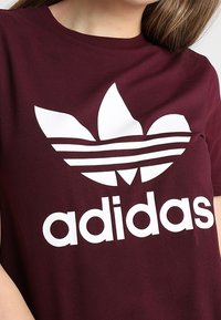 Acercamiento a una persona que lleva una camiseta marón de Adidas con un gran logo de trébol blanco y el nombre de la marca en el frente.