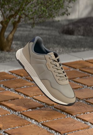 Zapatilla deportiva casual en beige y gris con suela blanca texturizada y cordones, descansando sobre un camino exterior de losas de madera y grava.