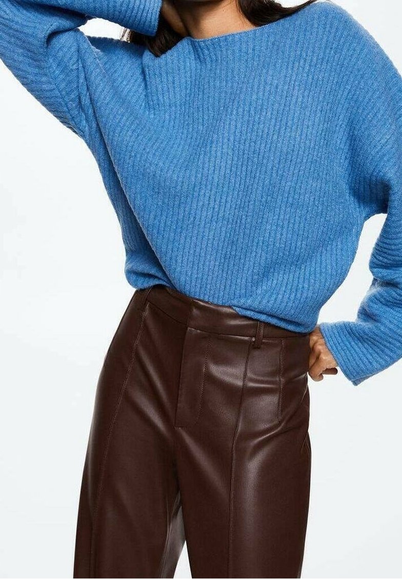 Frau in einem gerippten blauen Pullover und hochtaillierten braunen Lederhosen, die mit einem Arm hinter ihrem Kopf posiert.
