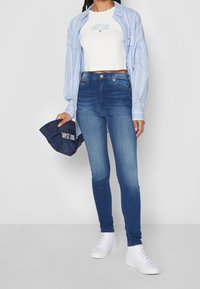 Vaqueros ajustados en azul medio con una textura suave, combinados con una camiseta blanca cortada y una camisa de rayas azul claro. Zapatillas blancas.