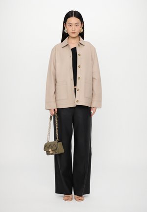 Bežinis mygtukais užsegamas švarkas su dviem priekinėmis kišenėmis, juodas trumpas viršus, juodos plačiakelnės kelnės ir tamsiai žalios rankinės su auksine grandine.