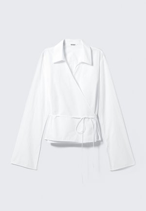 Valge ümar kaelusega pluus, mis on valmistatud siledast kangast. Tal on pikad varrukad ja sobitatud talje, millel on paela detail. Lihtne ja kaasaegne disain.