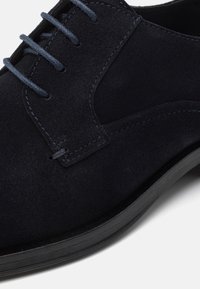 Gros plan sur une chaussure habillée pour homme en daim noir avec des lacets foncés et une semelle noire sur un fond clair.