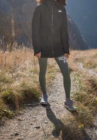 Giacca nera impermeabile con zip e cappuccio, leggings verdi con testo "NORTH" e sneakers grigie su un sentiero accidentato con erba.