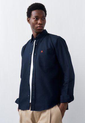 Νέος άντρας με μπλε σκούρο πουκάμισο πάνω από λευκό t-shirt και μπεζ παντελόνι, κοιτώντας προς τα πλάγια με τα χέρια στις τσέπες.