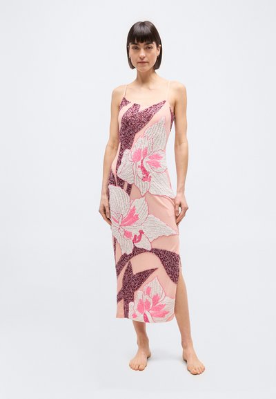 Γυναίκα που στέκεται ξυπόλητη, φορώντας ένα ροζ φόρεμα slip με μεγάλα υφασμένα λουλουδένια σχέδια σε λευκό και σκούρο ροζ, σε φόντο απλό και ανοιχτόχρωμο.