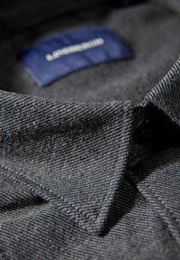 Donkergrijs denim overhemd met een close-up van de kraag, met een gestructureerde stof, stiksel details en een blauw merklabel aan de binnenkant.